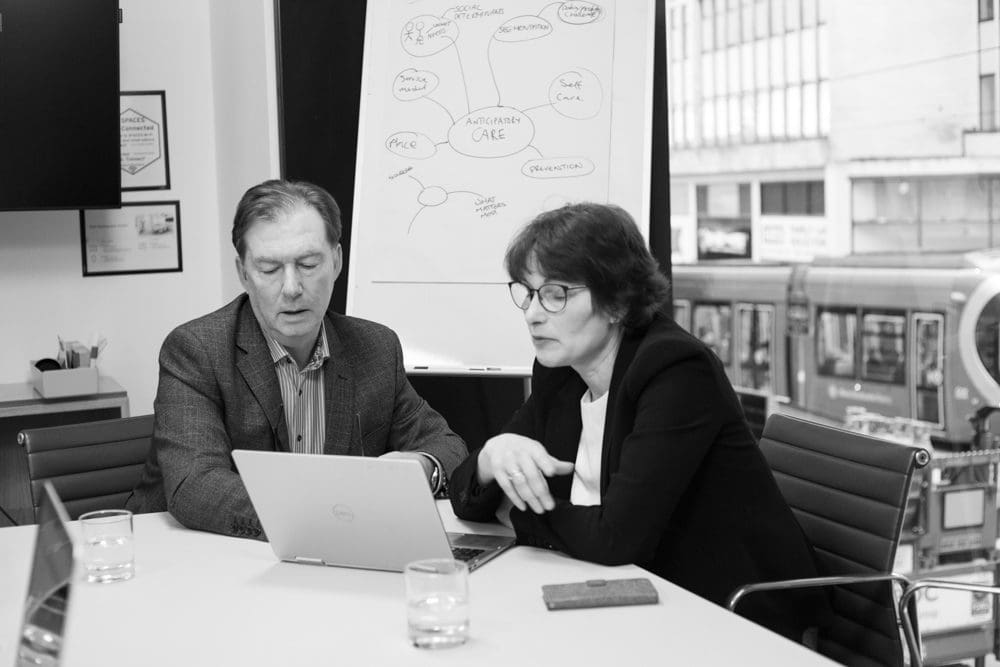 Two people in business attire sit at a table, looking at a laptop. A flipchart with a flowchart is behind them. Glasses of water and office supplies are on the table. Large windows show buildings and a tram outside, perfectly capturing the professional atmosphere through the lens of a Birmingham commercial photographer.