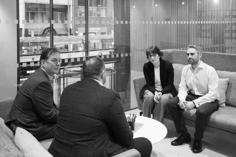 Four people in business attire sit on sofas around a small table in a modern office, engaged in a serious conversation. Captured by a Birmingham commercial photographer, the scene features glass walls and an urban view outside.
