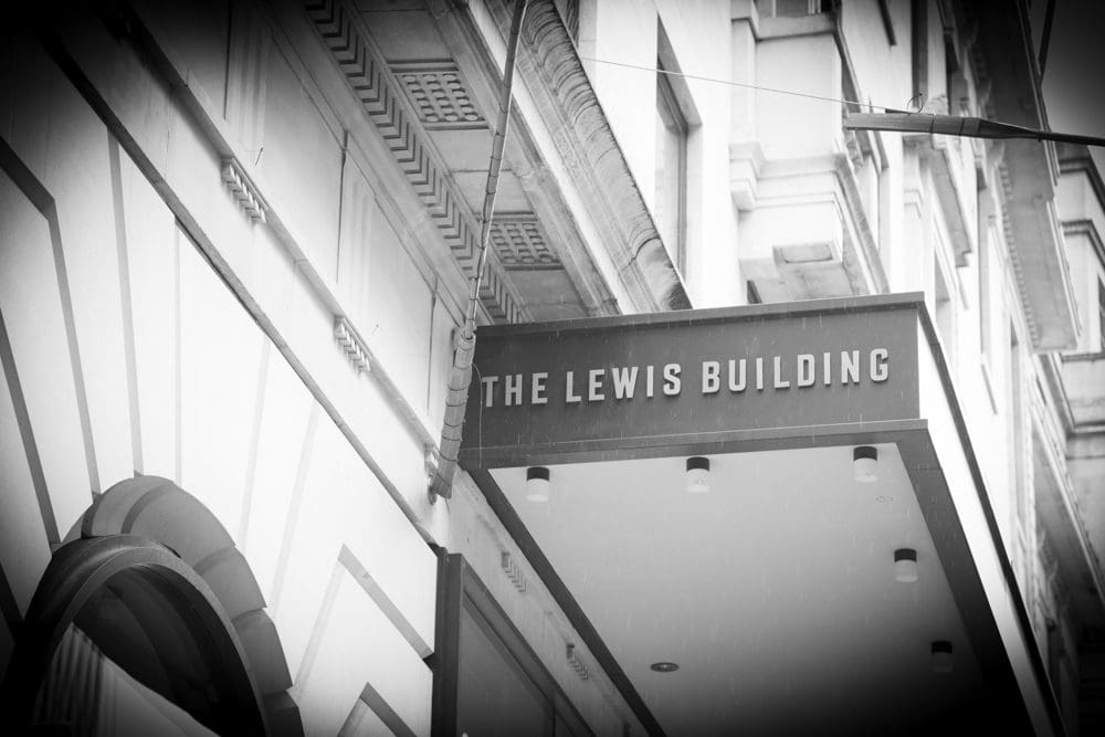 Black and white photo of a building entrance with a sign that reads THE LEWIS BUILDING. Captured by a Birmingham commercial photographer, the image highlights the sign mounted above a covered walkway attached to a classical-style façade.