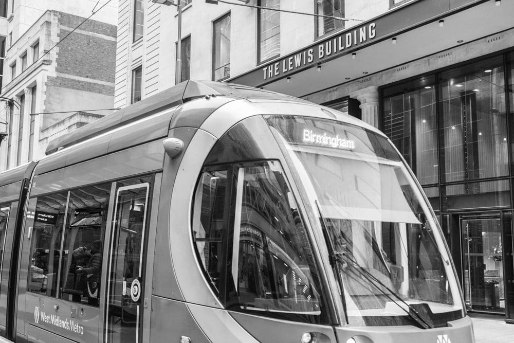 A modern tram labelled Birmingham travels through a city street in front of The Lewis Building, beautifully captured by a Birmingham commercial photographer. The image is in black and white.