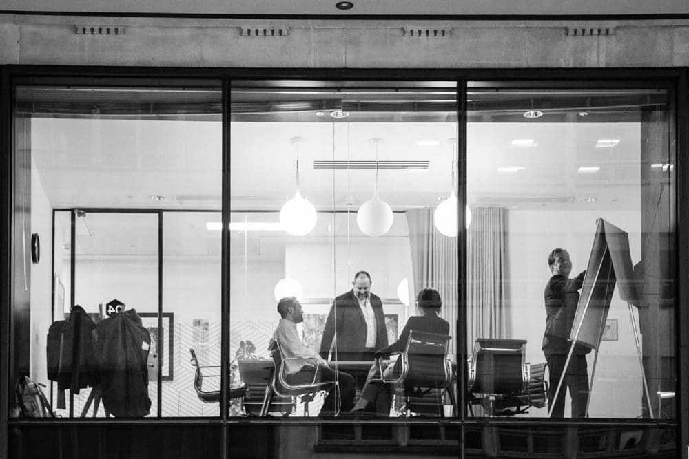 Four people in business attire are having a meeting in a modern office. Two are seated at a table, while two stand near a whiteboard. The scene, expertly captured by a Birmingham commercial photographer, is viewed through a large window. The image is in black and white.
