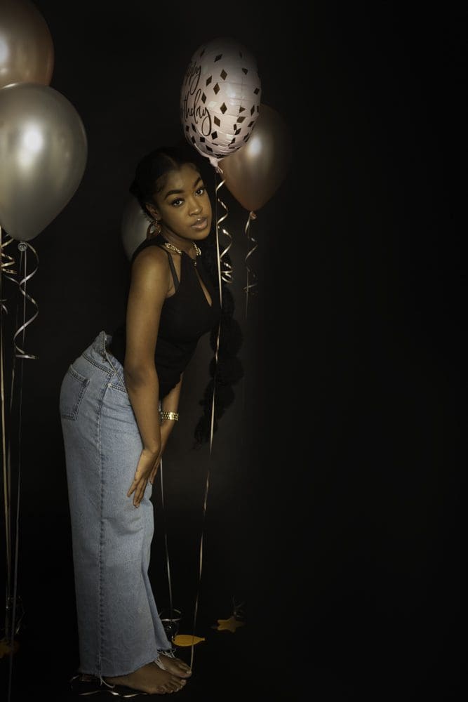 A woman in a black top and jeans poses barefoot with three metallic balloons against a dark background, looking towards the camera—an elegant moment captured by a Birmingham adult birthday photographer.