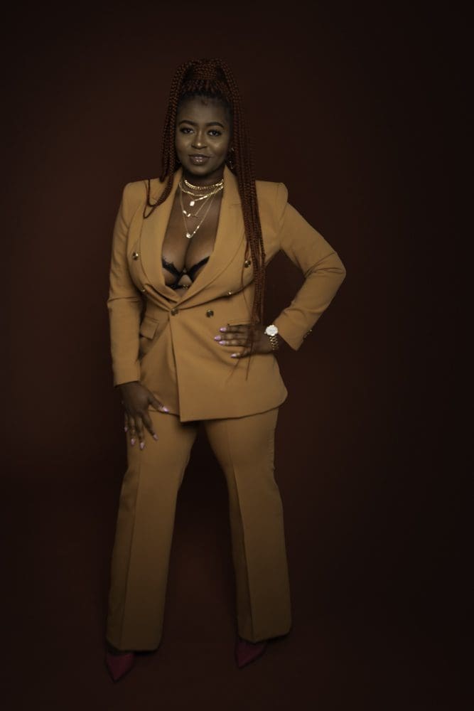 Woman stands confidently against a dark background, wearing a tan suit with a deep neckline, layered necklaces, and red shoes. She has long plaited hair and rests one hand on her hip, smiling slightly—a striking portrait captured by a Birmingham adult birthday photographer.