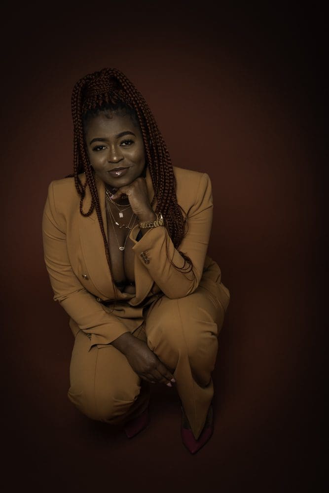 A woman with long plaits wears a tan suit and sits in a squatting pose, smiling softly with her chin resting on her hand against a dark brown background—an elegant portrait that showcases the artistry of a Birmingham adult birthday photographer.