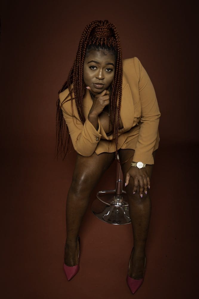 Woman with long plaits in a tan blazer and shorts sits on a stool, leaning forward with one hand under her chin, wearing pink heels, a watch, and purple nail varnish against a dark brown background—captured in an elegant portrait by a Birmingham adult birthday photographer.