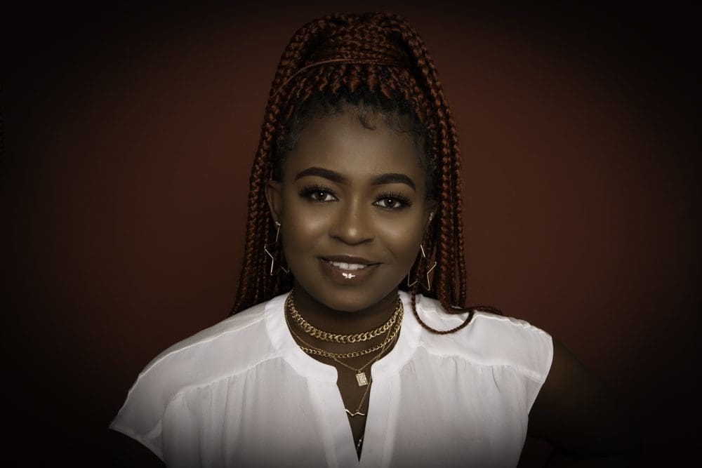 A woman with long, plaited reddish-brown hair smiles at the camera. She wears a white top, gold necklaces, and hoop earrings, with a dark reddish-brown background—captured in the signature style of a Birmingham adult birthday photographer.
