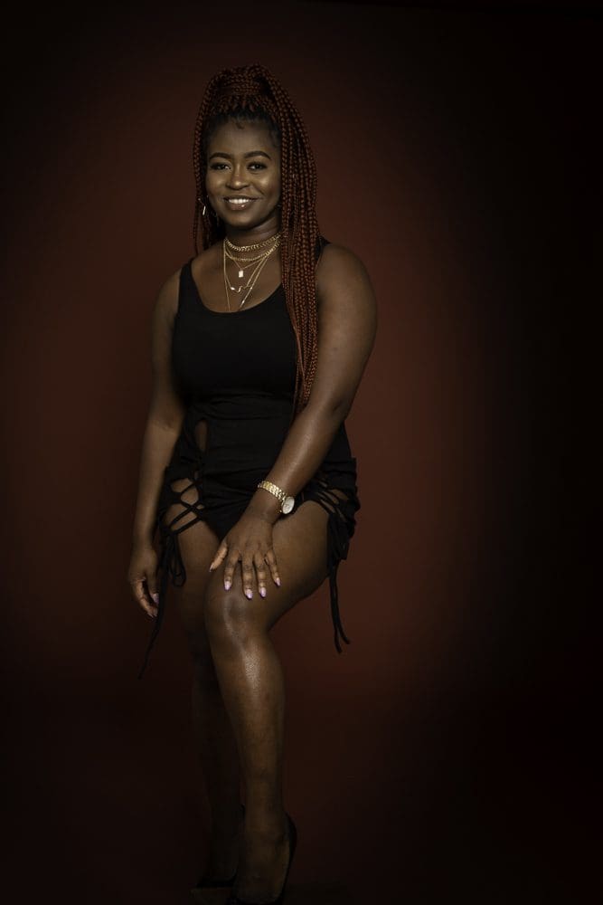 A woman with long plaited hair wears a black dress and layered necklaces, sitting with one knee raised, smiling against a dark brown background—perfectly capturing the elegant style celebrated by a Birmingham adult birthday photographer.