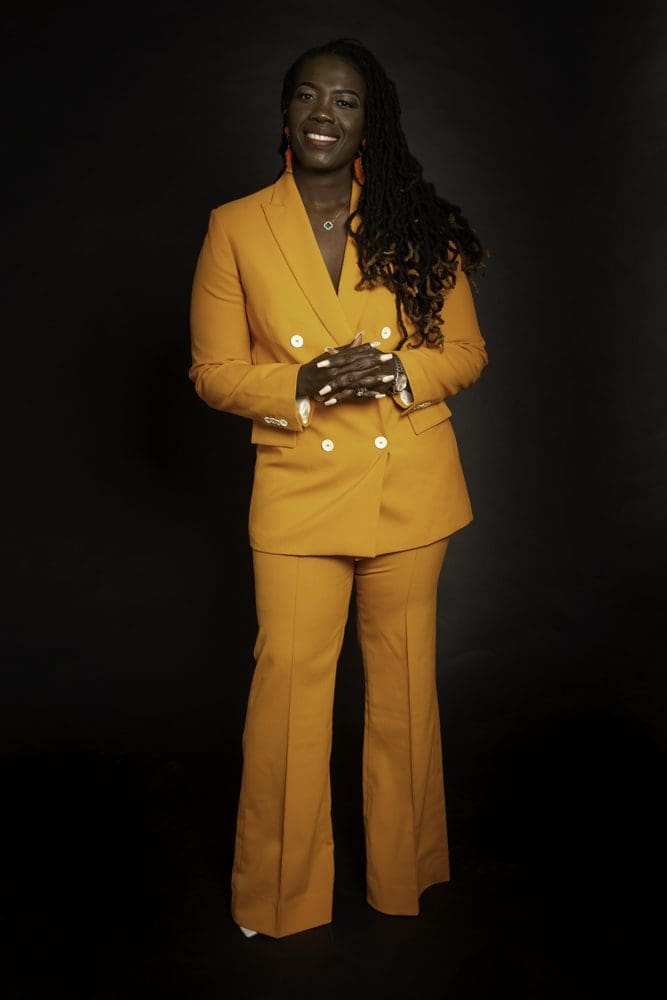 A person stands against a black background, smiling and wearing a bright orange suit with flared trousers. They have long plaited hair, orange earrings, and hands clasped in front—perfectly capturing the vibrant style that a Birmingham adult birthday photographer would love to highlight.