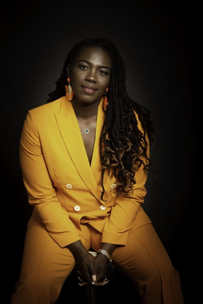 A woman with long, dark dreadlocks wears a bright orange suit and orange earrings, sitting against a dark background, looking confidently at the camera with her hands resting on her knees—captured in striking detail by a Birmingham adult birthday photographer.