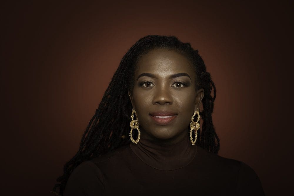 Woman with long dark plaited hair, wearing gold earrings and a dark polo neck, smiles softly against a dark maroon background—captured beautifully by a Birmingham adult birthday photographer.