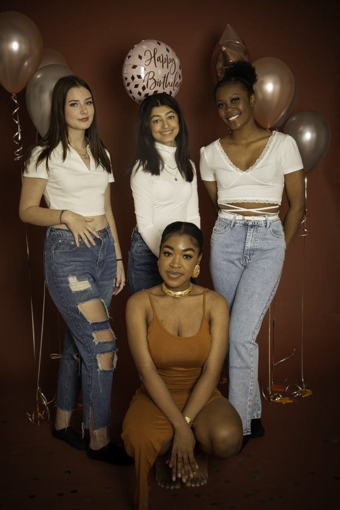 Four young women pose against a brown backdrop with birthday balloons, their smiles radiant as they celebrate together. Three stand wearing jeans and white tops, while one sits in front in a brown dress. Captured by a Birmingham adult birthday photographer, this joyful group portrait perfectly preserves the spirit of the occasion.
