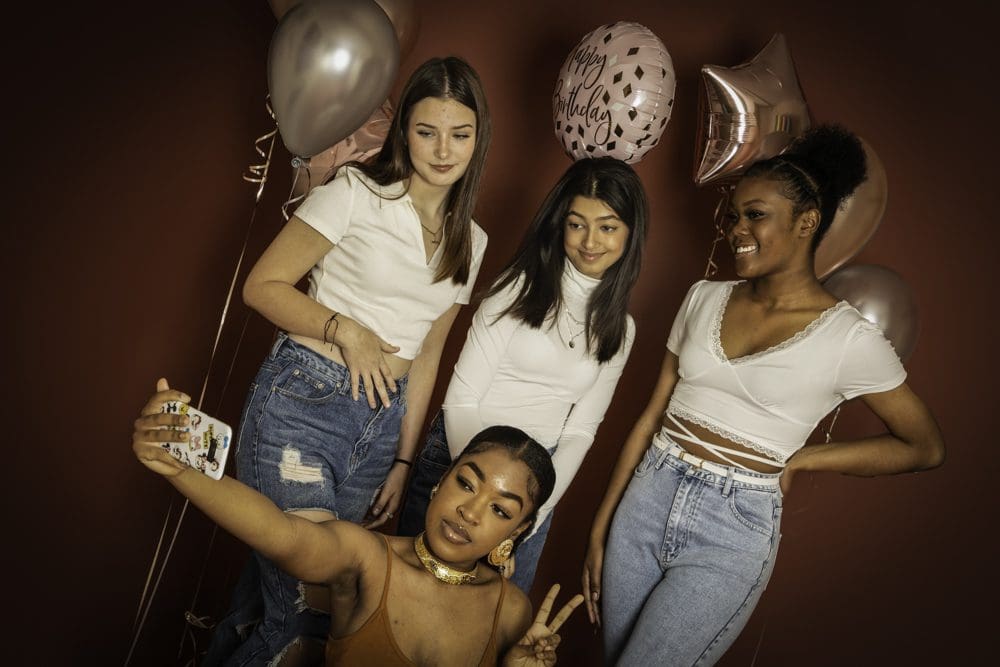 Four young women pose for a selfie at a party, standing in front of a red wall with metallic balloons, including one that says Happy Birthday. Three wear white tops and jeans, while the one taking the selfie wears a tan top. The joyful moment is perfectly captured by a Birmingham adult birthday photographer, ensuring memories from this special celebration last for years to come.