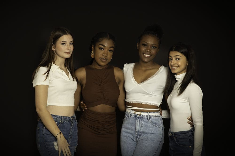 Four young women stand close together, smiling at the camera. They wear casual outfits, including jeans, skirts, and crop tops, against a plain dark background—capturing the fun and energy of a celebration through the lens of a Birmingham adult birthday photographer.