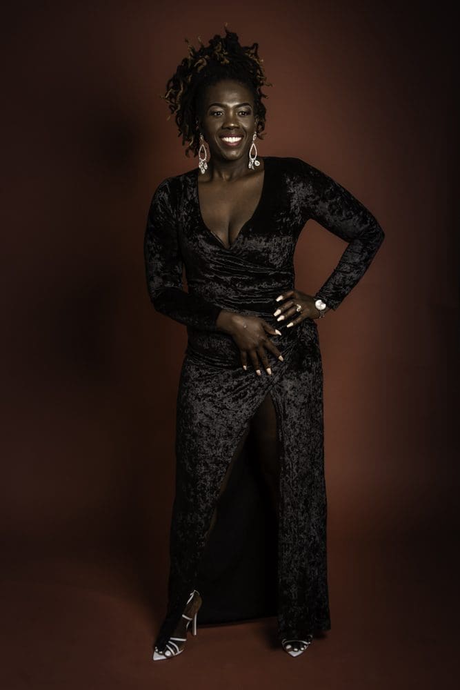 A person smiles confidently, wearing a long black velvet dress with a deep neckline and high slit, silver earrings, and heels, posing against a dark brown background—a perfect portrait for anyone seeking a Birmingham adult birthday photographer to capture stylish milestone moments.