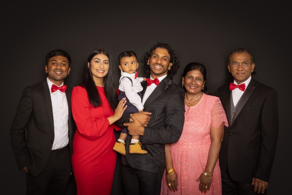 Six people pose together against a black background, dressed in formal attire with red accents. A man holds a baby, with three men in suits and bow ties, and two women in elegant dresses smiling at the camera.