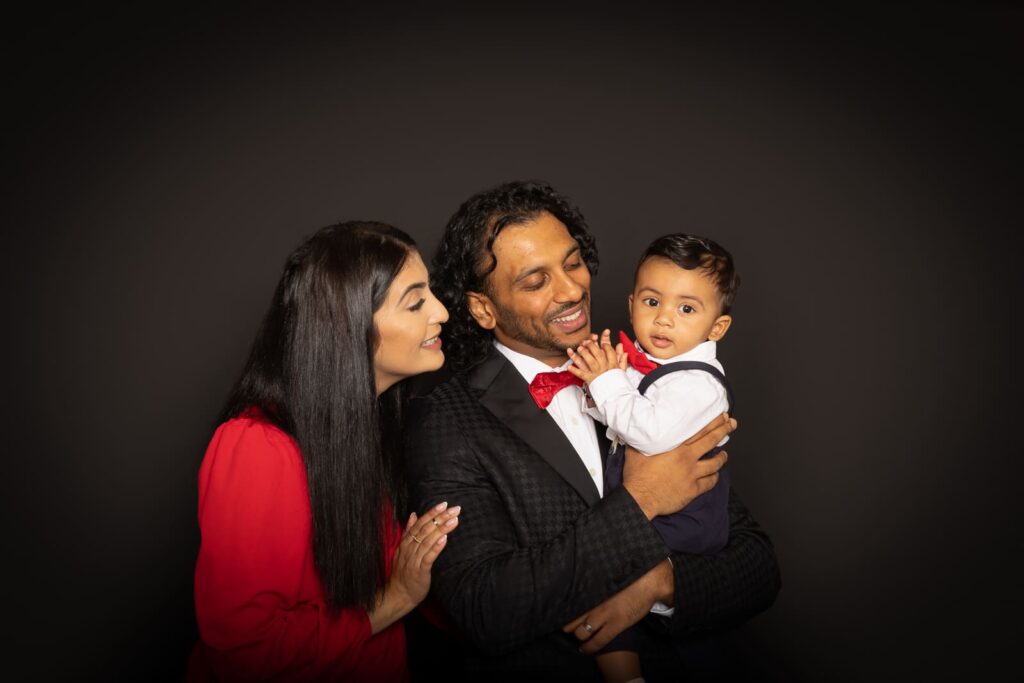 A smiling family of three poses together, with a woman in red, a man in a dark suit with a red bow tie holding a young child, all against a plain dark background.