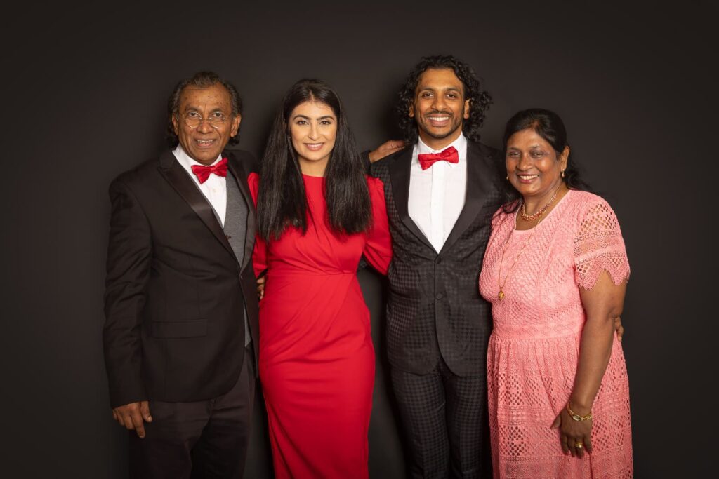 Four people stand together smiling, dressed formally. Two men wear black suits with red bow ties, one woman wears a red dress, and the other wears a pink lace dress. The background is plain and dark.
