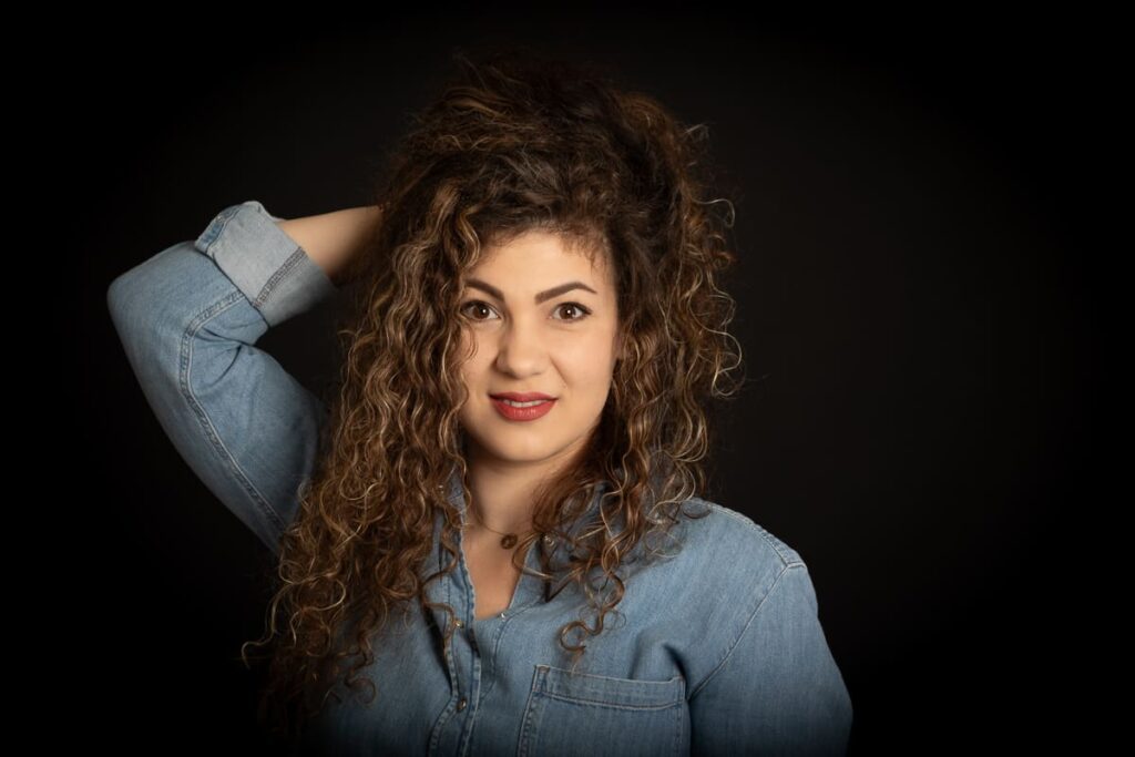 Woman with long curly hair wearing a denim shirt poses against a black background, one hand touching her hair, and looks confidently at the camera—an elegant image captured by a skilled Birmingham portrait photographer.