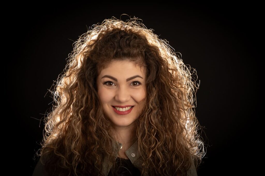 Woman with long, curly hair smiling at the camera against a black background, wearing a grey shirt and minimal jewellery. Captured by a skilled Birmingham portrait photographer, her face is well-lit, highlighting her features and hair.