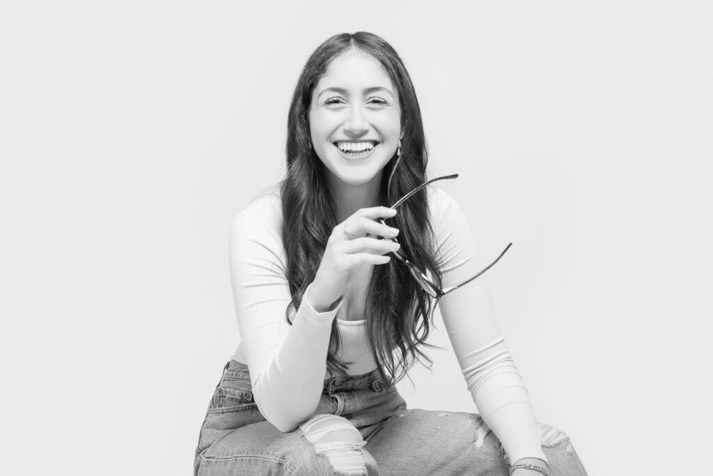 A smiling woman with long hair sits in casual ripped jeans and a long-sleeve shirt, holding spectacles in one hand. Captured by a skilled Birmingham portrait photographer, the black and white photo features a light background, highlighting her relaxed and cheerful demeanour.