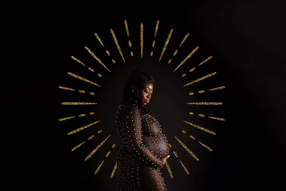 A pregnant woman in a sheer, spotted dress holds her belly, standing against a dark background with golden rays radiating out behind her, symbolising light or celebration.