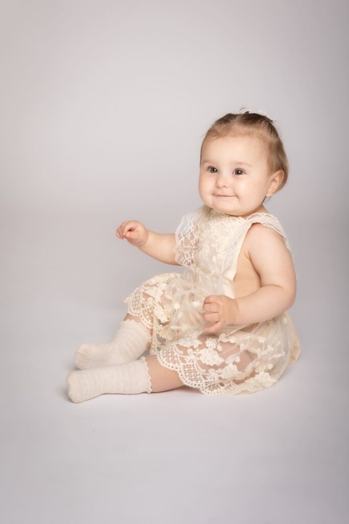 A baby sits on the floor wearing a cream-coloured lace dress and white socks, smiling and looking slightly to the side against a plain light background.