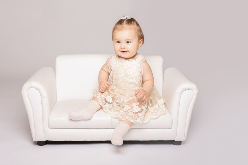 A smiling baby wearing a cream lace dress and white socks sits on a small white sofa against a light background.
