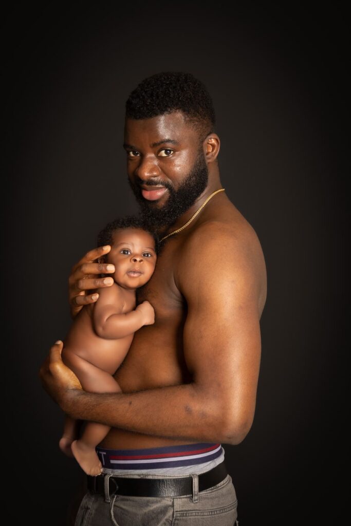 A bare-chested man with a beard holds a smiling, naked baby close to his chest. Both are looking at the camera against a plain dark background.