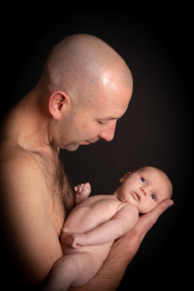A shirtless bald man gently cradles a naked baby in his arms against a black background, gazing lovingly at the infant.