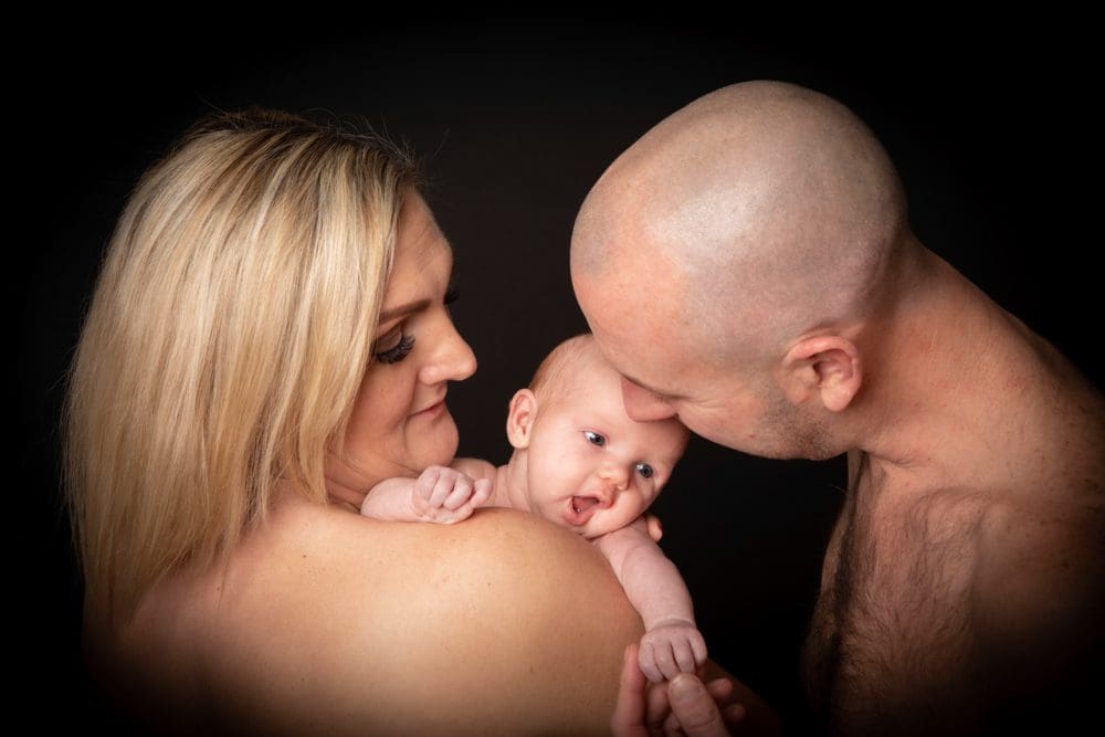 A woman holds a baby while a man gently kisses the baby's head. All three are topless and facing each other, with a dark background creating a soft, intimate atmosphere.