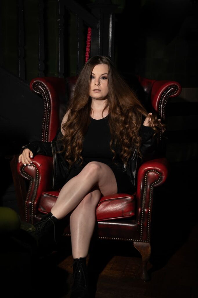A woman with long brown hair wearing a black dress sits confidently on a red leather armchair, legs crossed, in a dimly lit room—captured in striking detail by a Birmingham portrait photographer.