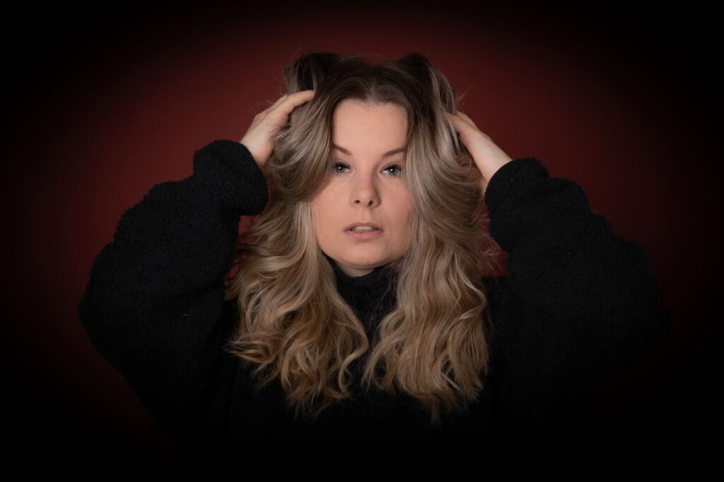 Woman with long wavy blonde hair wearing a black jumper, holding her hair up with both hands, looking directly at the camera against a dark red background—an elegant composition captured by a Birmingham portrait photographer.