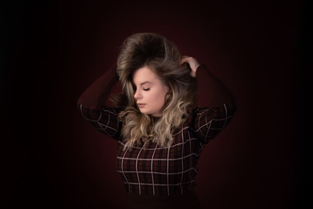 A woman with long, wavy blonde hair poses with her eyes closed, wearing a checked, long-sleeved shirt against a dark background. Captured by a Birmingham portrait photographer, her hands are in her hair, creating an evocative and stylish image.