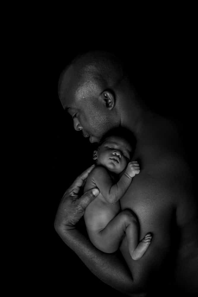 A man gently holds a sleeping newborn baby against his bare chest. The image is in black and white with a dark background, highlighting the peaceful and intimate moment between parent and child.