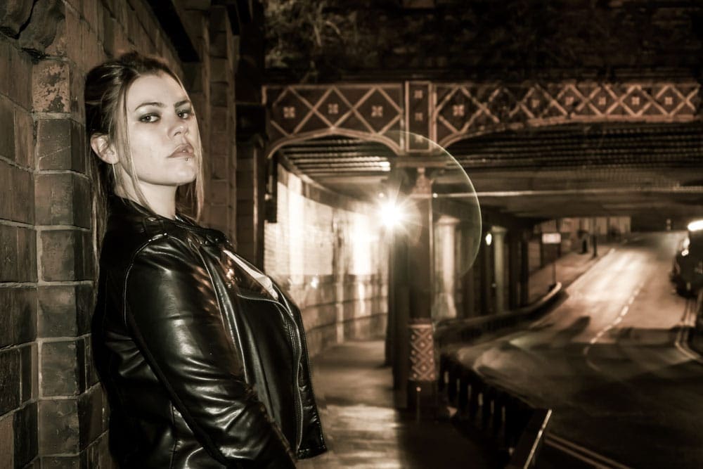 A woman in a leather jacket leans against a brick wall at night, with a dimly lit street and ornate bridge in the background.