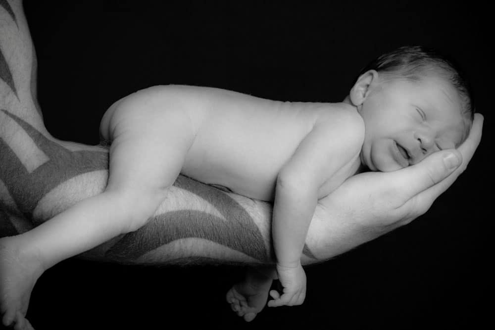A newborn baby sleeps peacefully on an adult’s tattooed arm and hand, supported gently in a horizontal position against a black background—an intimate moment beautifully captured by a Birmingham newborn photographer.