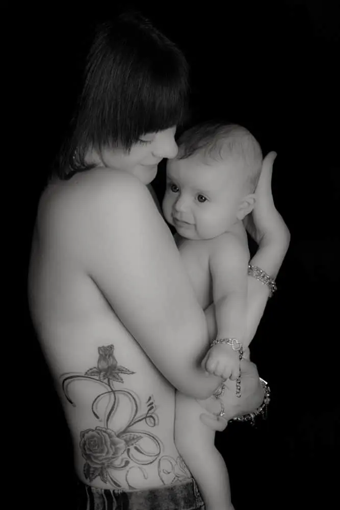 A tattooed woman holds a smiling baby against her chest. Both are topless and appear happy. The image is black and white, with a dark background highlighting their close bond.