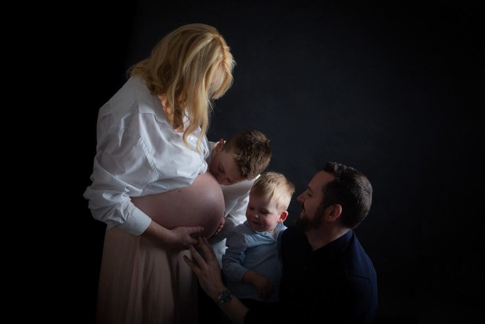 A pregnant woman stands with a young boy gently kissing her bump, while a man and a toddler sit beside them, touching her bump and looking up lovingly. The background is dark, highlighting the family.