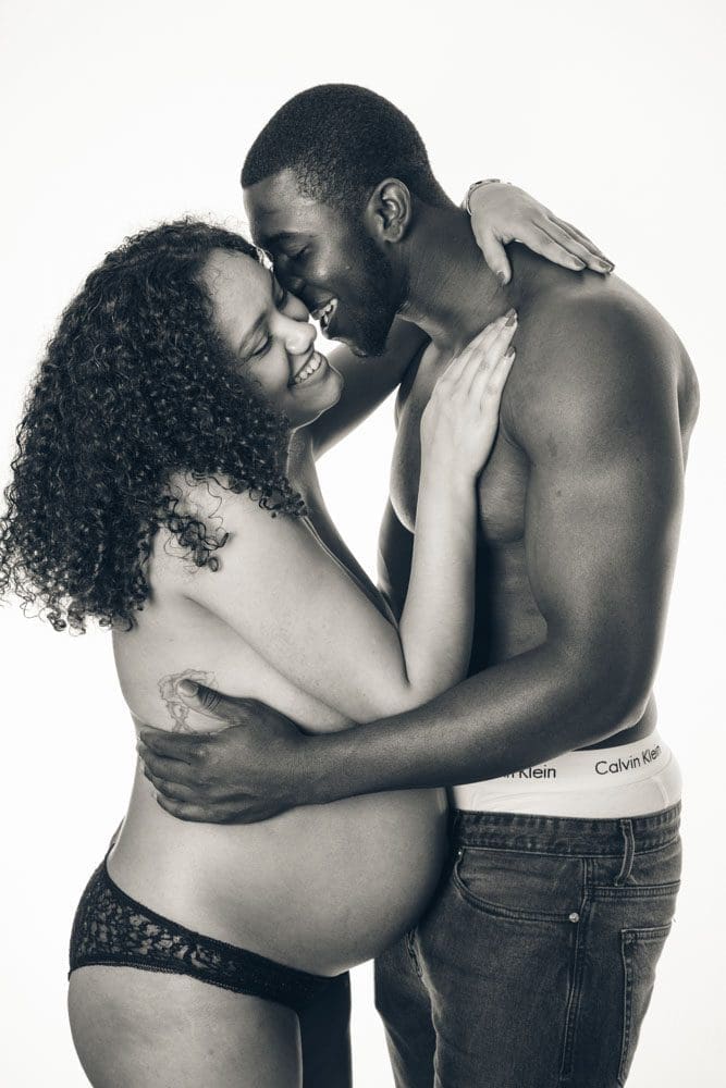 A pregnant woman and a man embrace and smile, both partially dressed, sharing an intimate, joyful moment against a plain background.