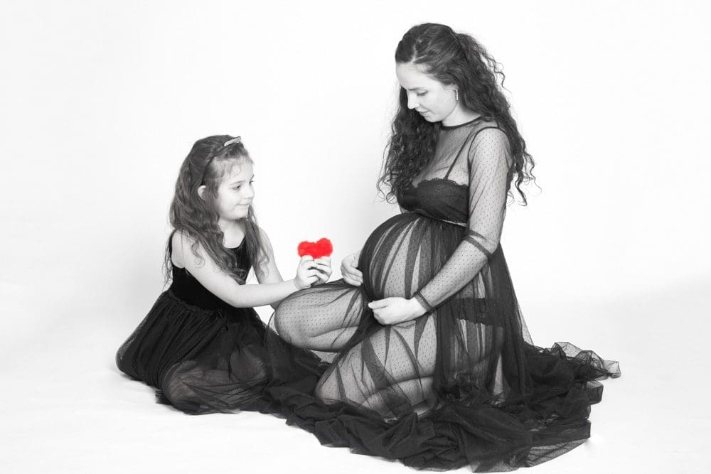 A young girl in a black dress hands a red flower to a pregnant woman in a sheer black dress. Both are sitting on the floor against a white background.