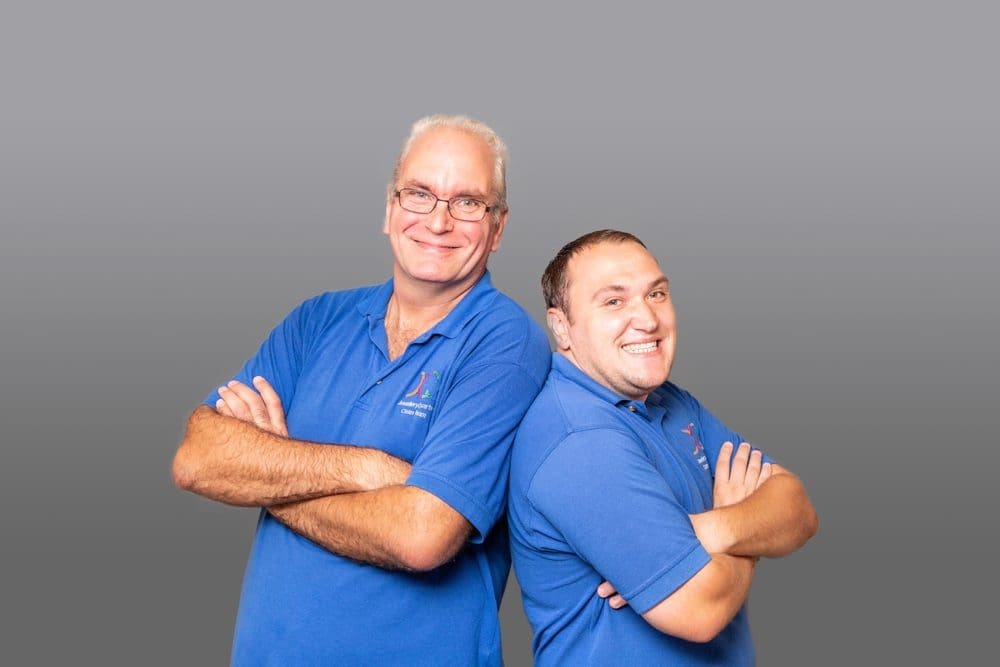Two men in blue polo shirts stand back-to-back with arms crossed, smiling at the camera against a plain grey background—captured with a professional touch by a Birmingham commercial photographer.