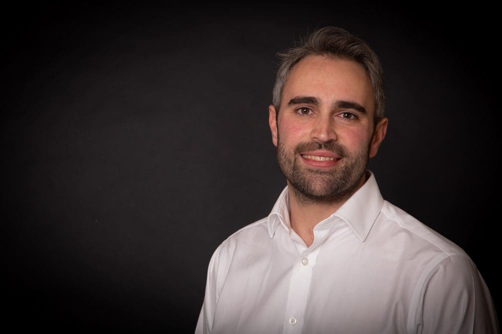 A man with short dark hair and beard, wearing a white dress shirt, smiles at the camera against a dark background in this striking portrait by a Birmingham commercial photographer.