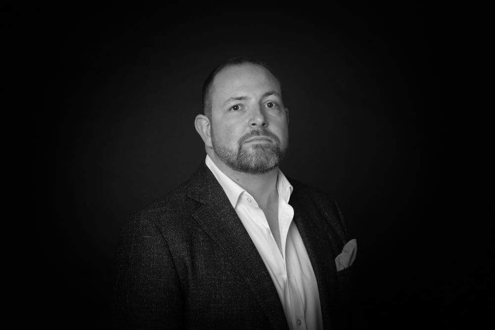 Black and white portrait of a man with short hair and a beard, wearing a suit jacket, white shirt, and pocket square, posing against a dark background—captured with the distinctive style of a Birmingham commercial photographer.