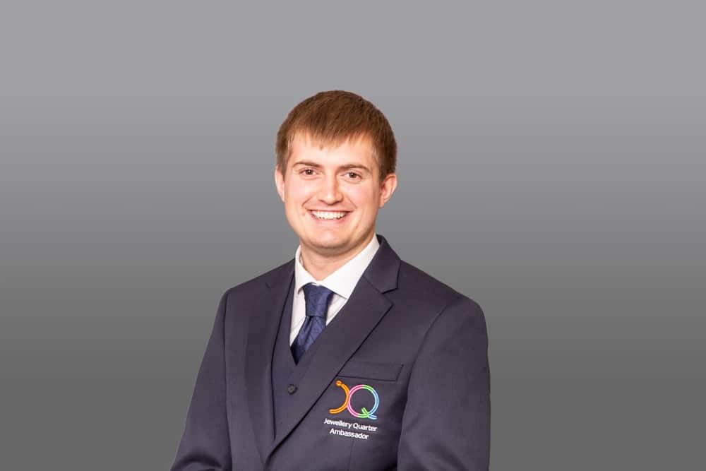 A young man in a dark suit and tie stands against a grey background, smiling. Captured with the polished eye of a Birmingham commercial photographer, his jacket features a colourful logo and the words “Q Ambassador” embroidered on it.