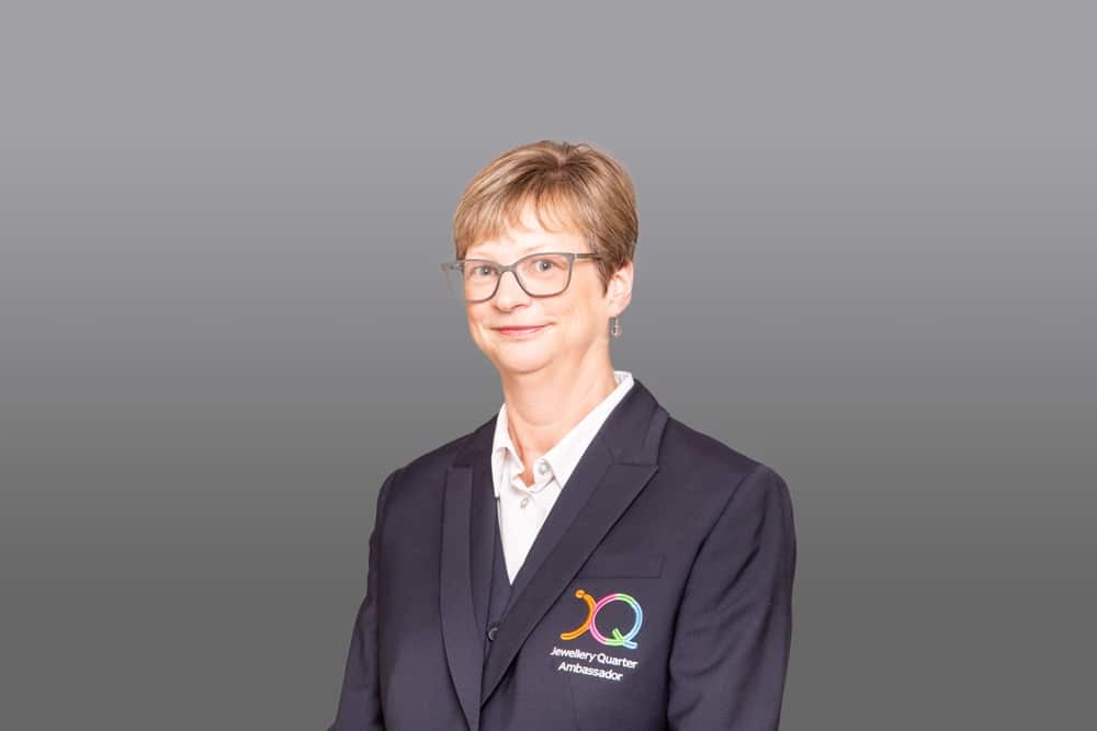 A person with short fair hair and glasses, wearing a dark blazer with a colourful logo and Jewellery Quarter Ambassador embroidered on it, stands in front of a plain grey background—captured in the distinctive style of a Birmingham commercial photographer.