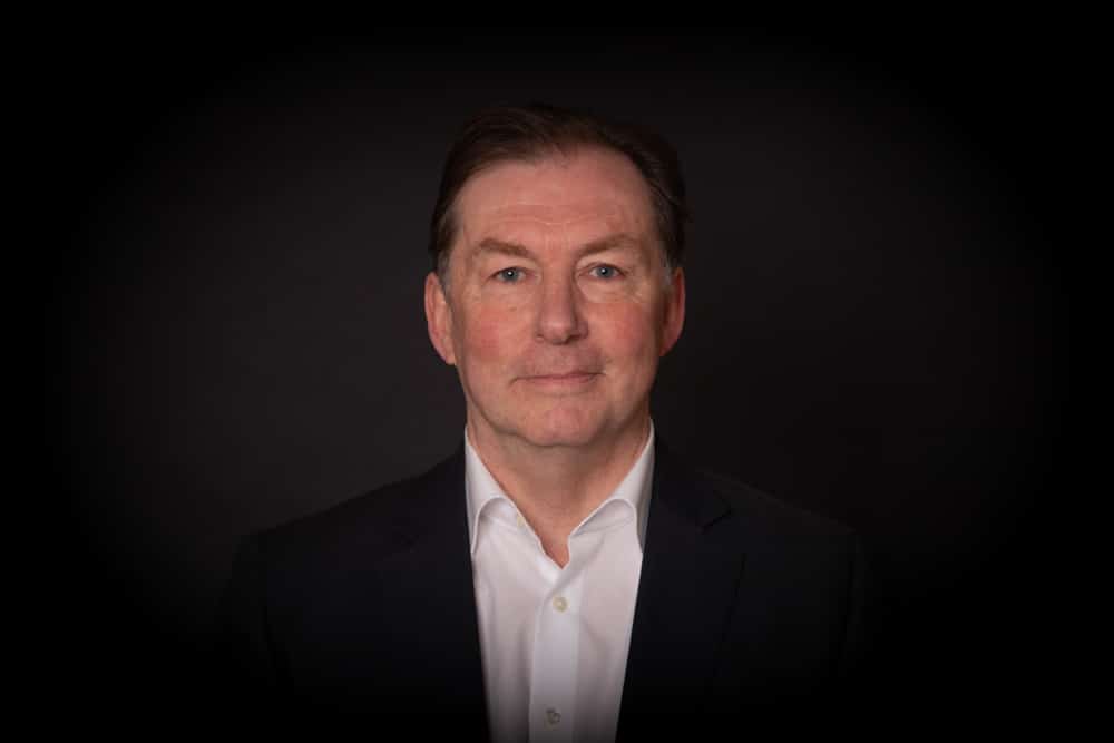 A middle-aged man with short brown hair, wearing a white shirt and dark blazer, poses against a plain black background, looking directly at the camera with a neutral expression—captured in the polished style of a Birmingham commercial photographer.