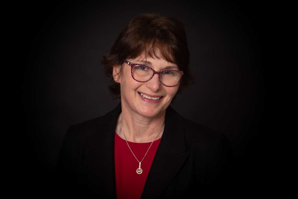 A woman with short brown hair, wearing glasses, a black blazer, a red top, and a necklace, smiles against a dark background in this striking portrait by a Birmingham commercial photographer.