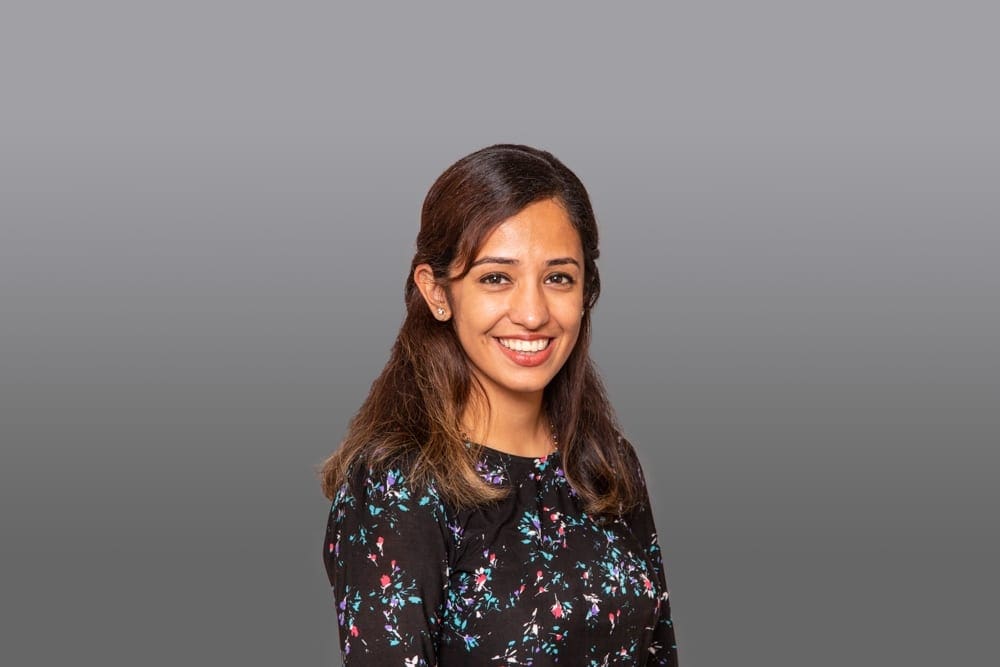 A woman with long brown hair, wearing a black floral top, smiles whilst standing in front of a plain grey background in a portrait captured by a Birmingham commercial photographer.