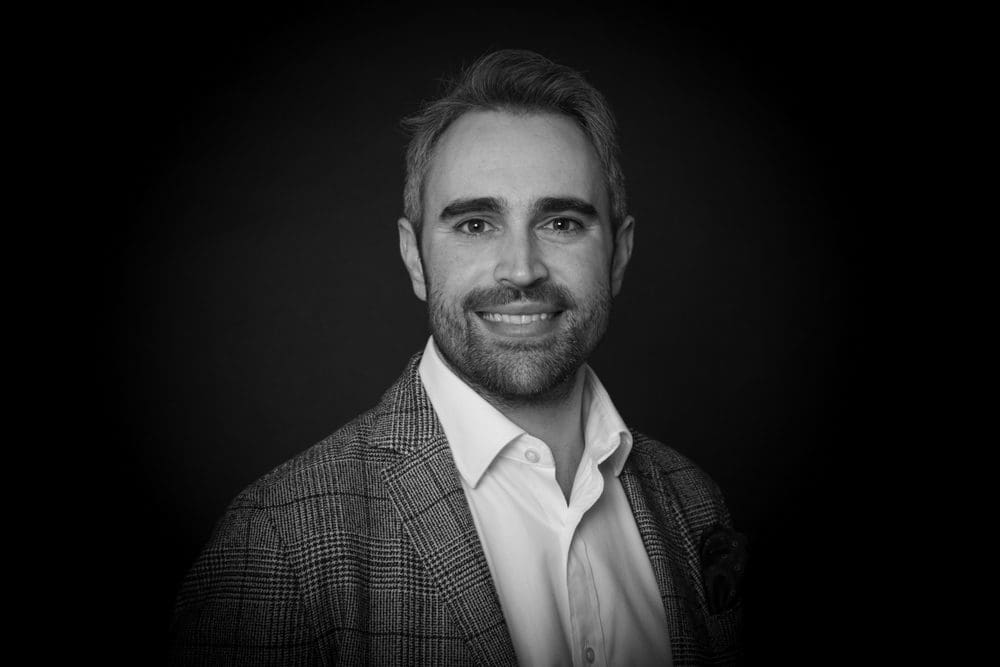 Black and white portrait of a man with short hair and a beard, wearing a checked blazer and white shirt, smiling at the camera against a dark background—captured by a skilled Birmingham commercial photographer.