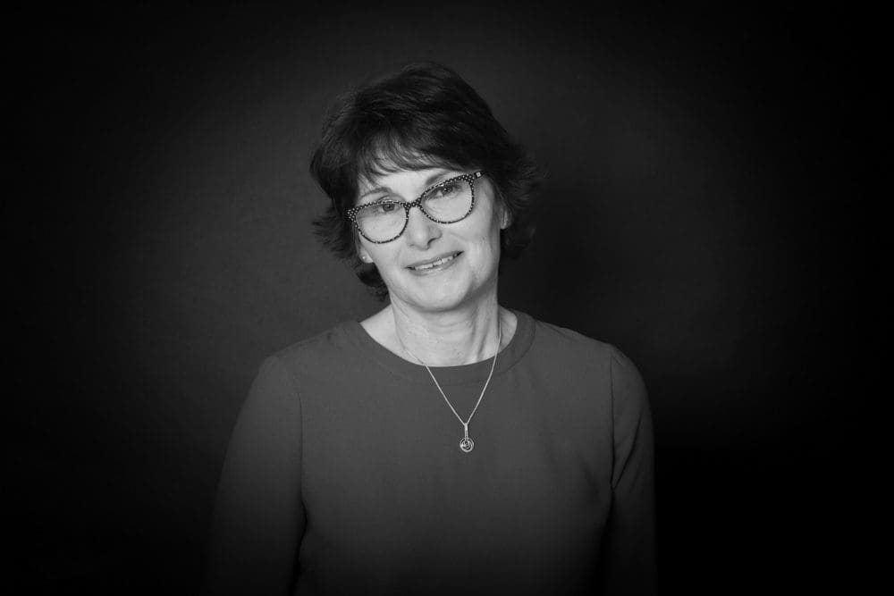 Black and white portrait by a Birmingham commercial photographer of a woman with short hair and glasses, wearing a pendant necklace and a plain top, standing against a dark background and smiling softly.